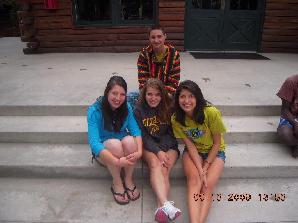 Four young women sitting on concrete steps in front of a wooden building with a dark green double door, smiling at the camera, with a young man standing behind them, also smiling.