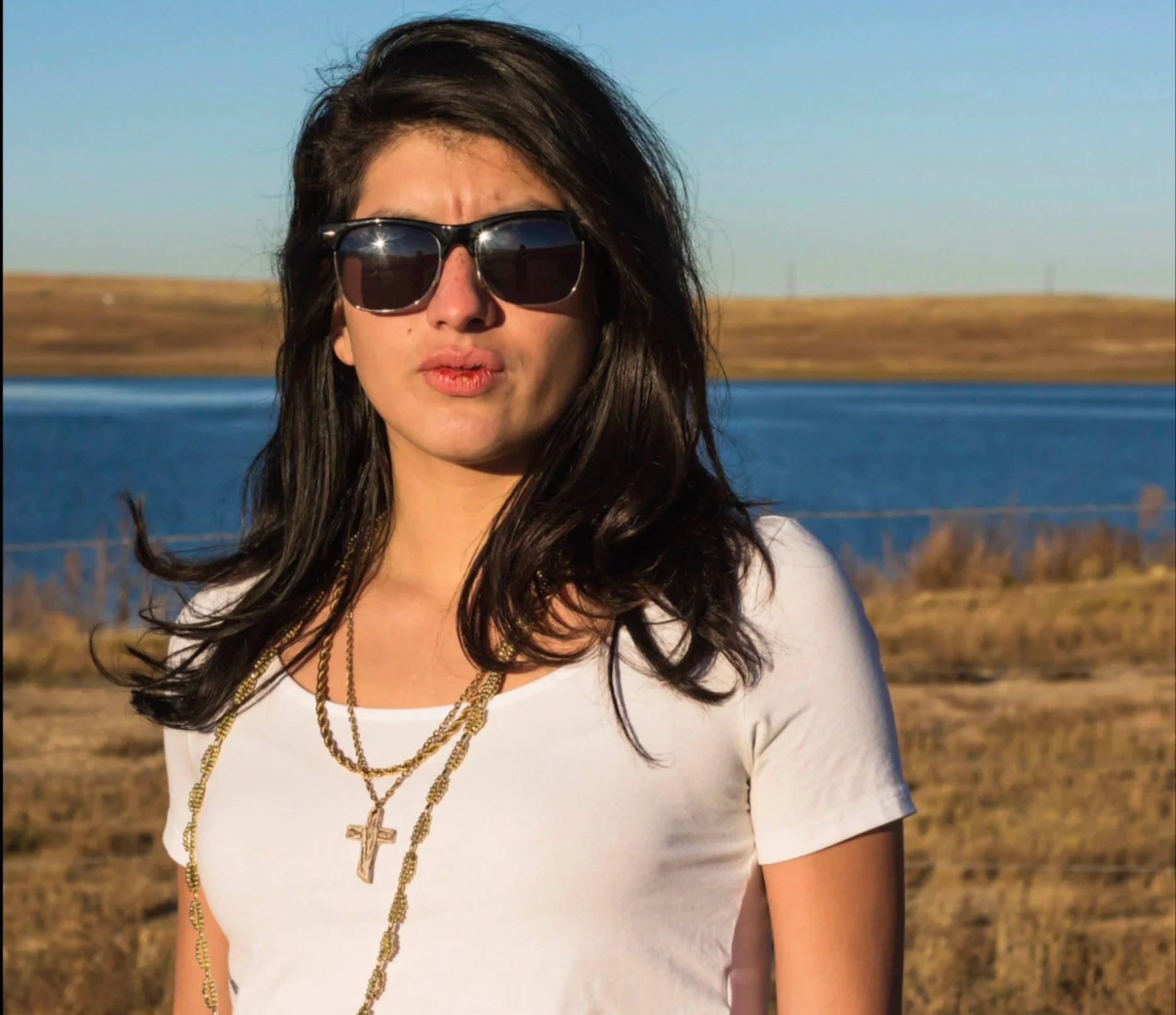 A woman with dark wavy hair wearing large black sunglasses, a white T-shirt, and multiple gold necklaces, standing outdoors with a body of water and a clear sky in the background.