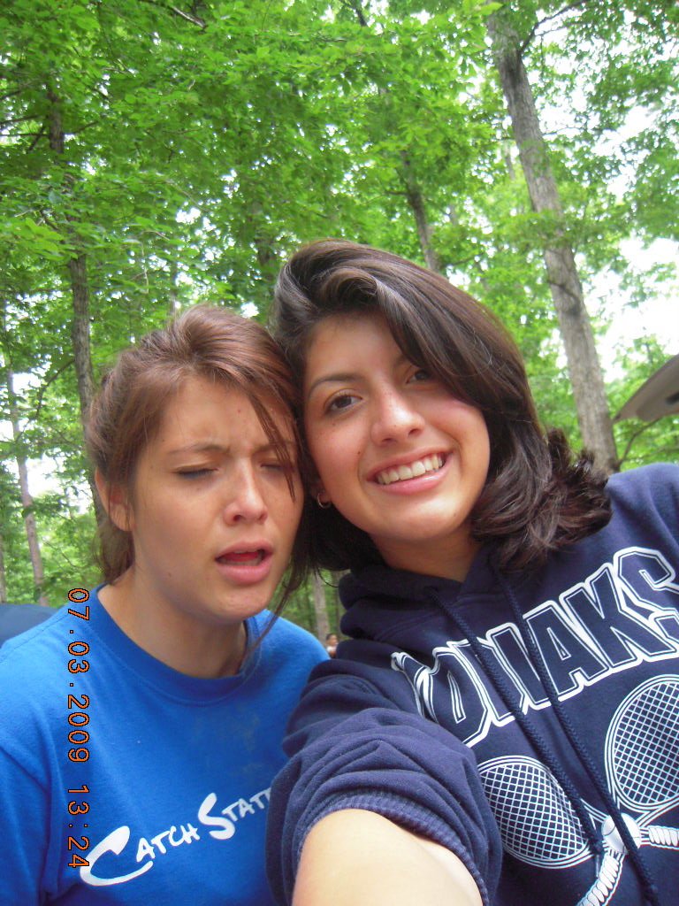 Two young women taking a selfie outdoors in a wooded area. One has light brown hair and a distressed expression, wearing a blue shirt. The other has dark brown hair, smiling, wearing a navy hoodie. Both are surrounded by tall trees with green leaves.