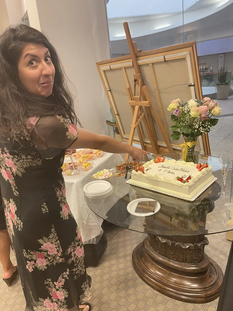 Woman in floral dress pointing at a cake with the text "Let's Celebrate More" on a glass table with plates, flowers, and decorations in the background.