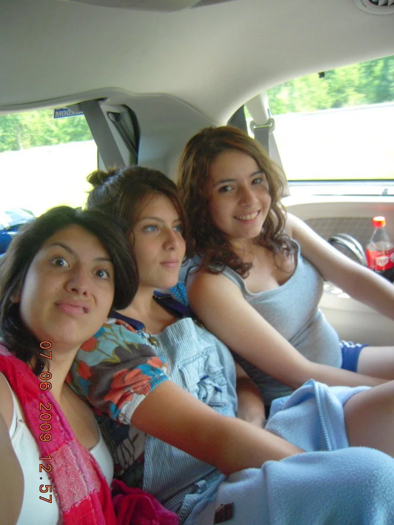 Three young women sitting inside a car, smiling and looking at the camera with a blurred green landscape outside the window.