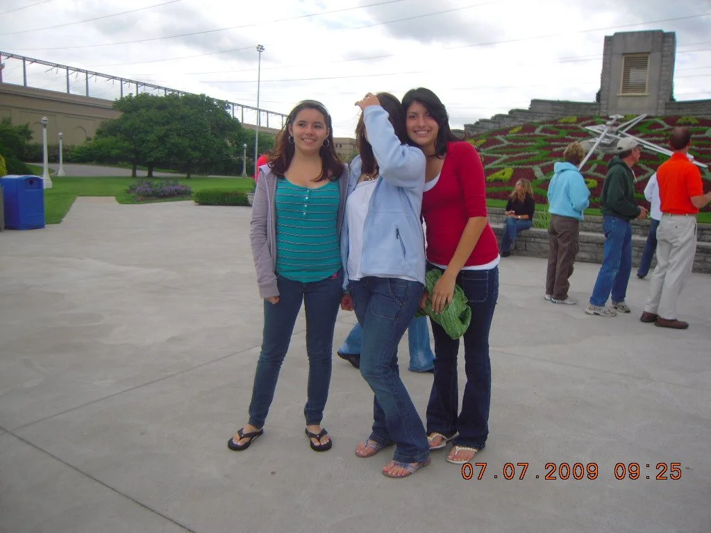 Three young women standing on a concrete pathway at a park, smiling, with others in the background near a large floral display and a sculpture, on a cloudy day.