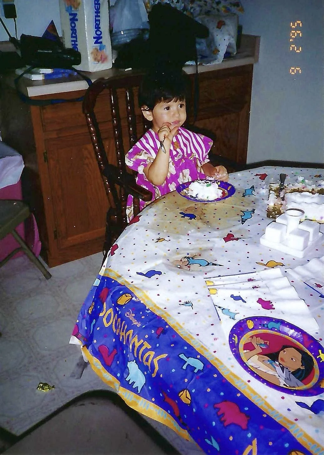 A young girl in a pink striped dress is sitting at a table with a birthday cake and a plate of cake with white frosting. The tablecloth has a colorful party hat and birthday theme with characters on it. The girl is holding a fork and eating. The back