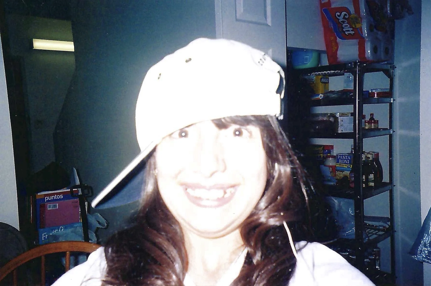 A woman with long brown hair wearing a white baseball cap and white top, smiling at the camera. The background shows a kitchen or pantry with shelves containing food items like pasta and snacks.