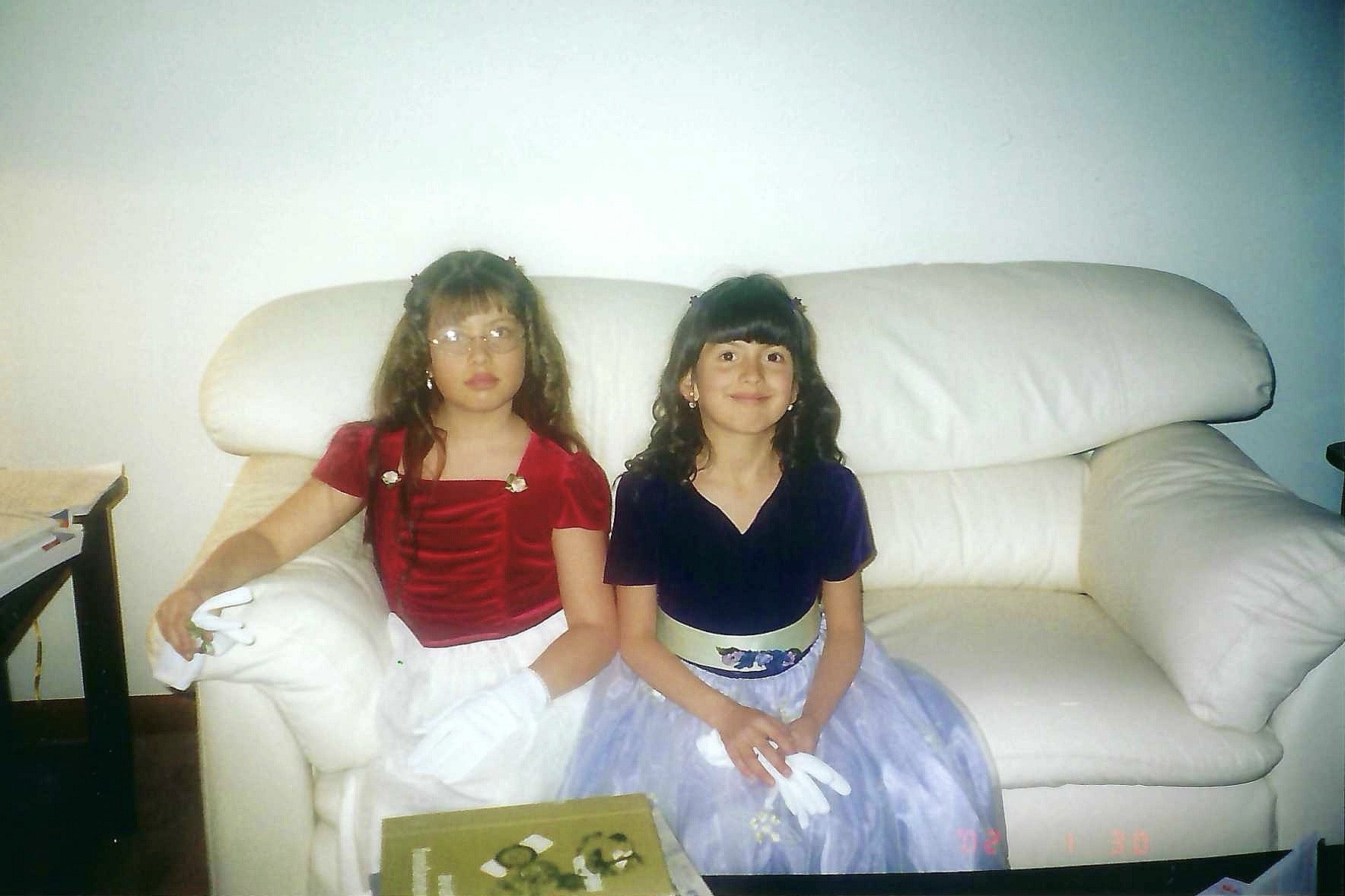 Two young girls sitting on a cream-colored sofa, one in a red top and white skirt, the other in a purple top and lavender skirt, in a living room with a table in front and plain white walls.