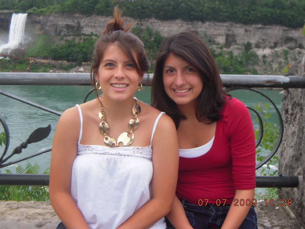 Two women smiling near a body of water with a waterfall in the background, standing in front of a metal railing.