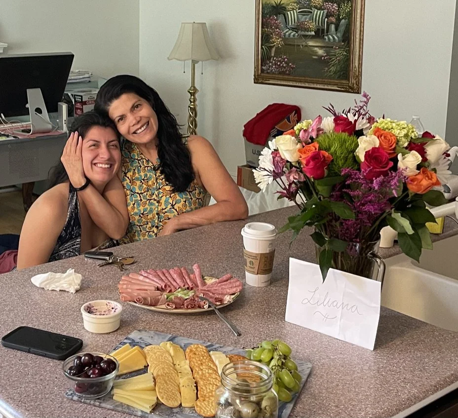 Two women sitting at a kitchen counter, smiling and leaning into each other. The counter has a bouquet of multicolored roses, a plate of assorted meats and cheese, grapes, and a coffee cup. A handwritten sign with the name 'Liliana' is in front of th