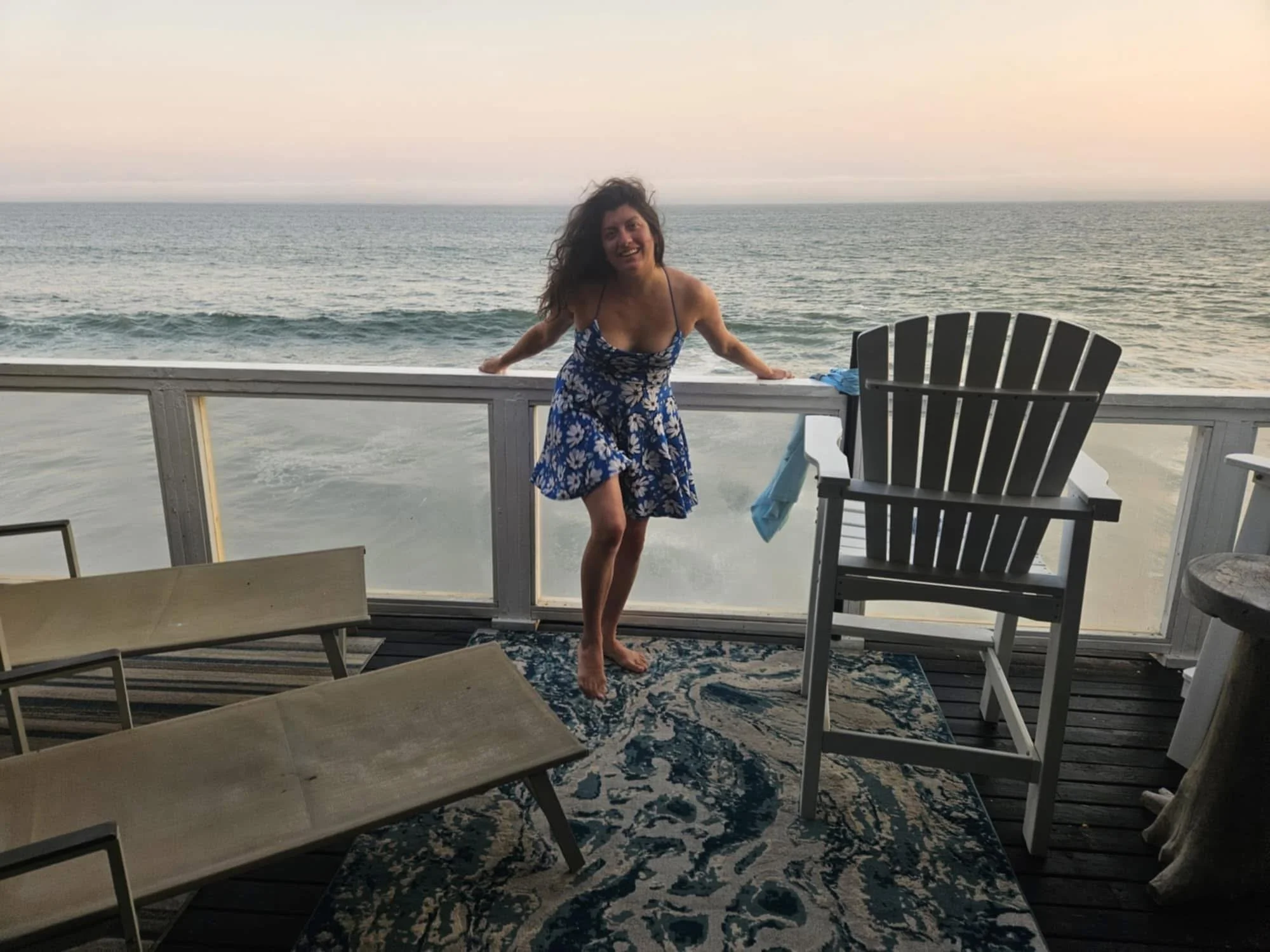 A woman in a floral blue dress smiling on a balcony overlooking the ocean at sunset. There are empty chairs and a rug on the wooden deck.