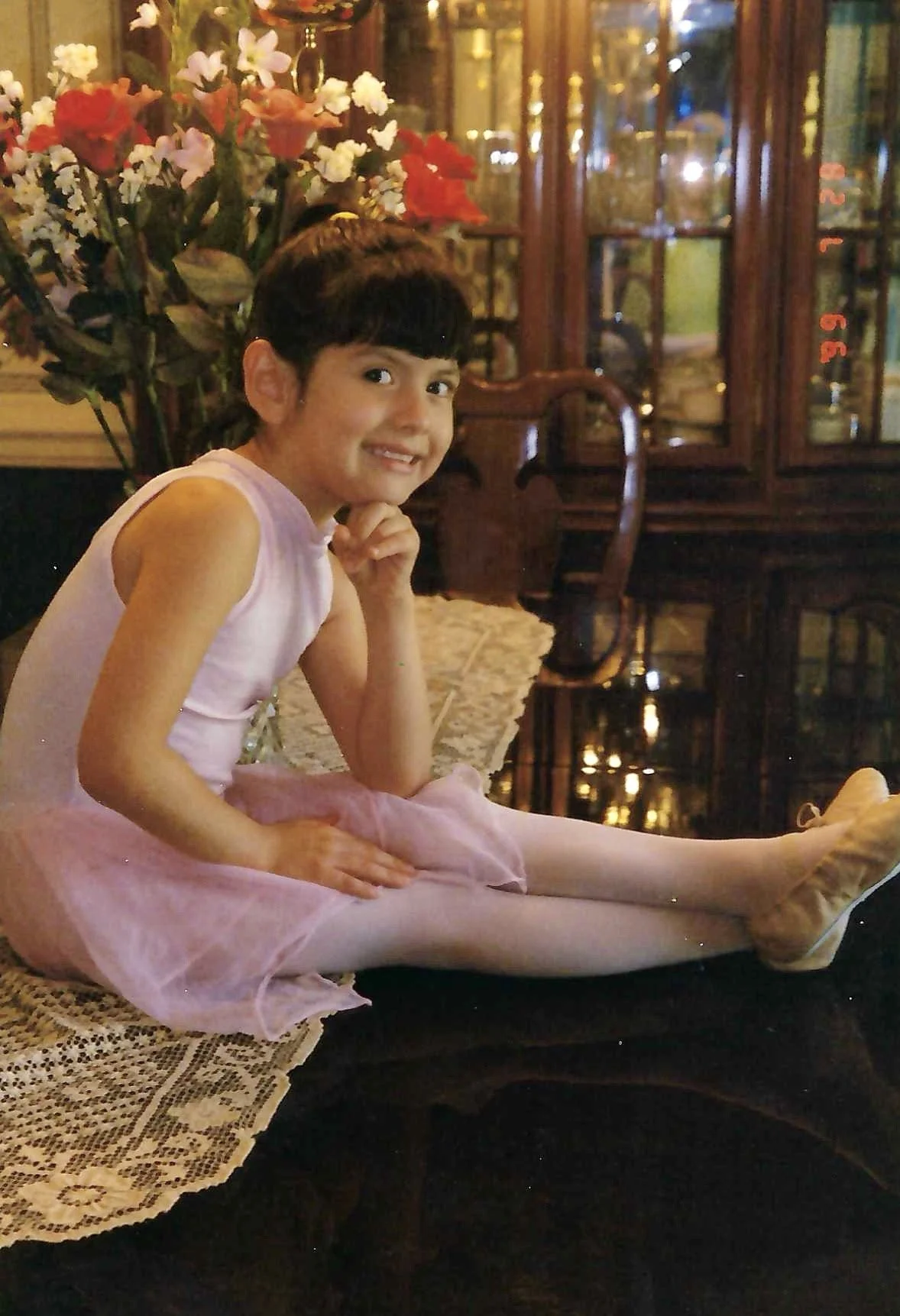 A young girl in ballet attire sitting on a lace table runner in a dimly lit room with wooden furniture, a bouquet of white, red, and pink flowers in the background, and a glass cabinet with decorative items.