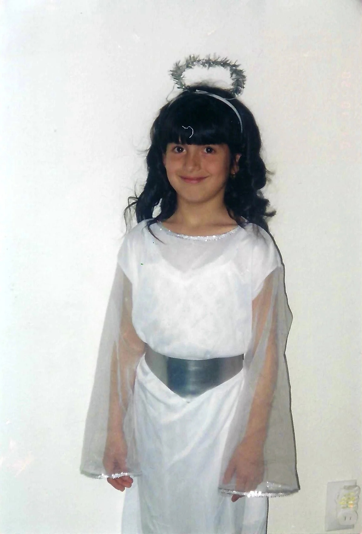 A young girl dressed as an angel, wearing a white gown with silver accents, a black belt, and a halo headband with tinsel, standing against a plain white wall.