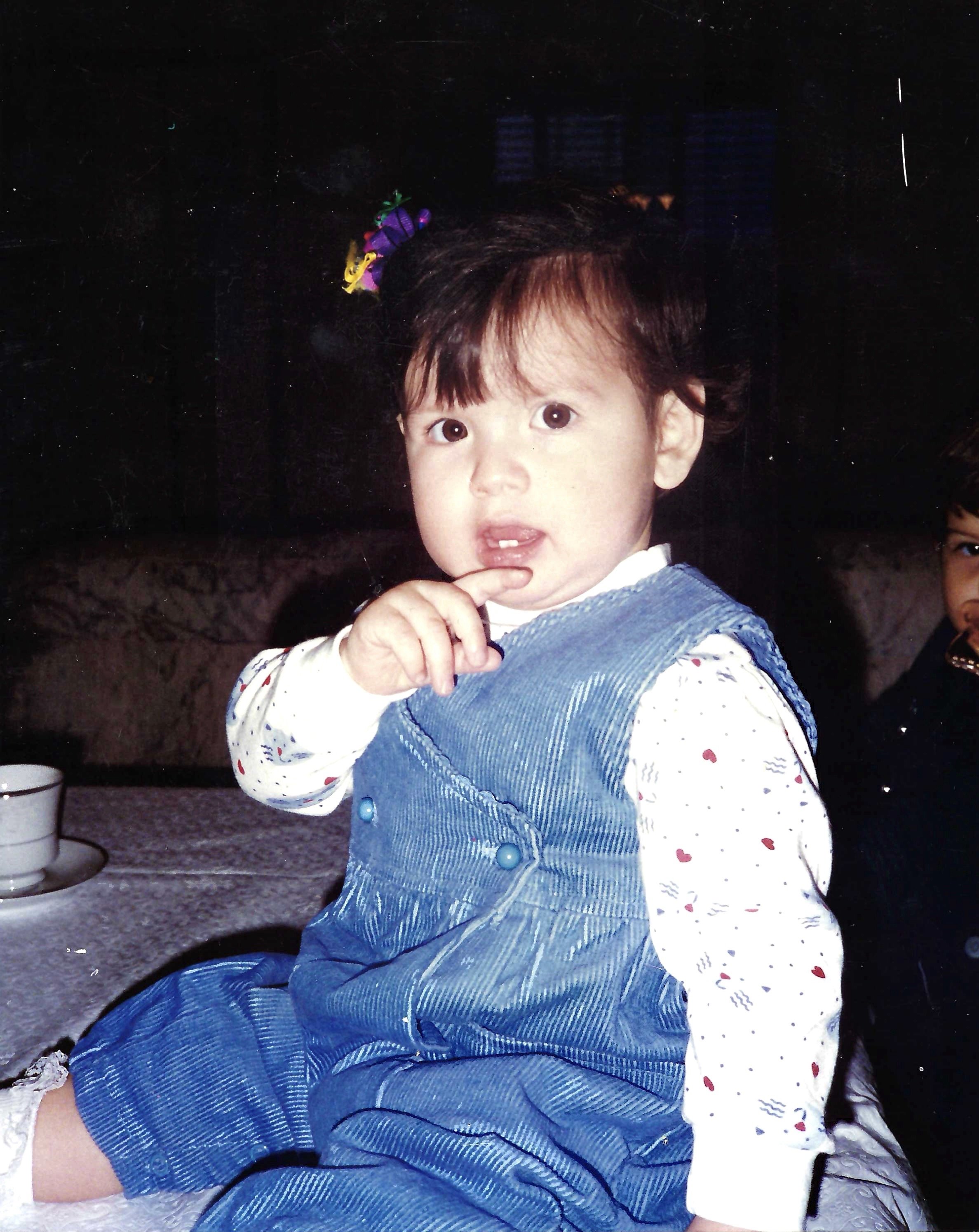 Young girl with brown hair and big eyes, wearing a white shirt with red hearts and blue overalls with large buttons, posing indoors with her finger near her lips.