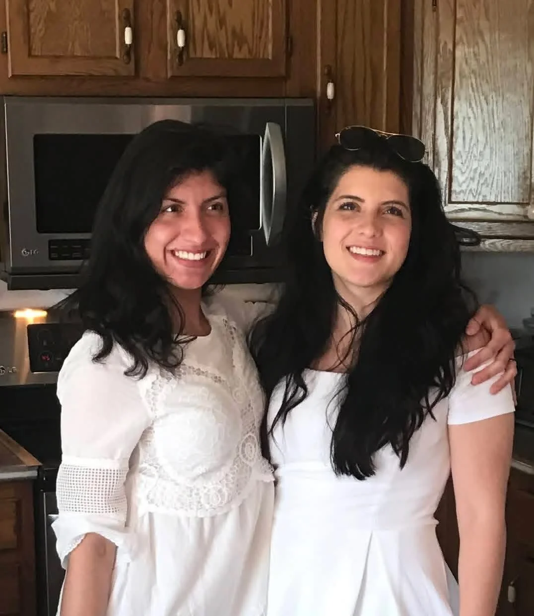 Two women with dark hair and white dresses smiling and hugging in a kitchen