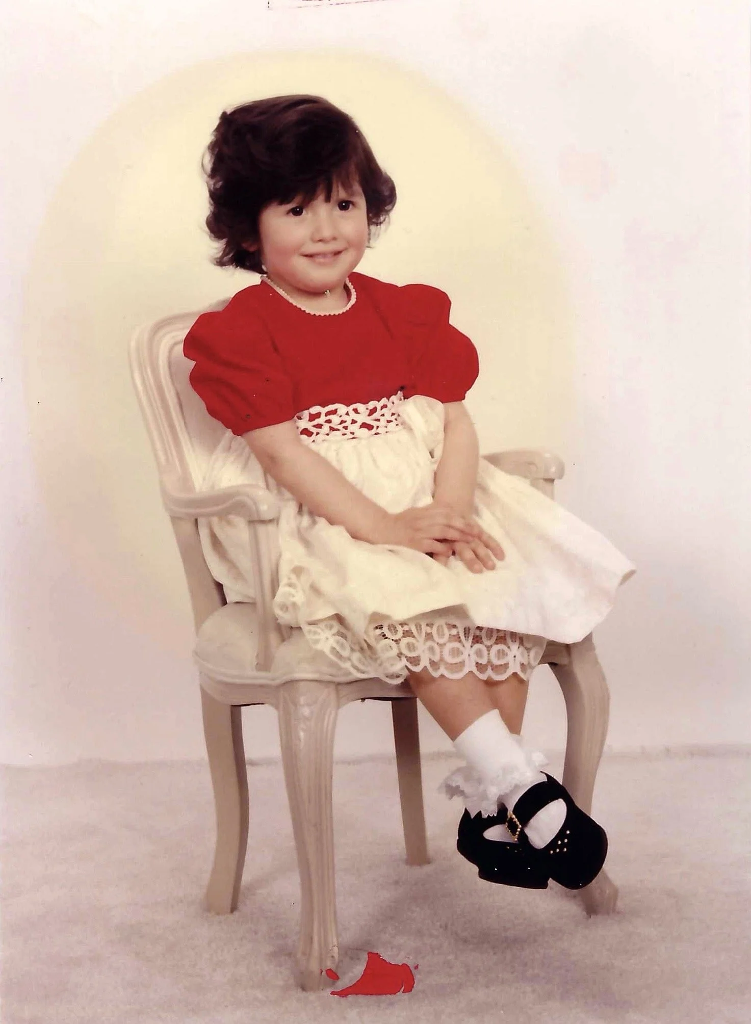 A young girl with short, dark curly hair sitting on a vintage wooden chair in a white room. She is wearing a red top with puffed sleeves and a pearl necklace, a cream-colored skirt with lace, white socks, and black shoes. Red fabric is on the floor u