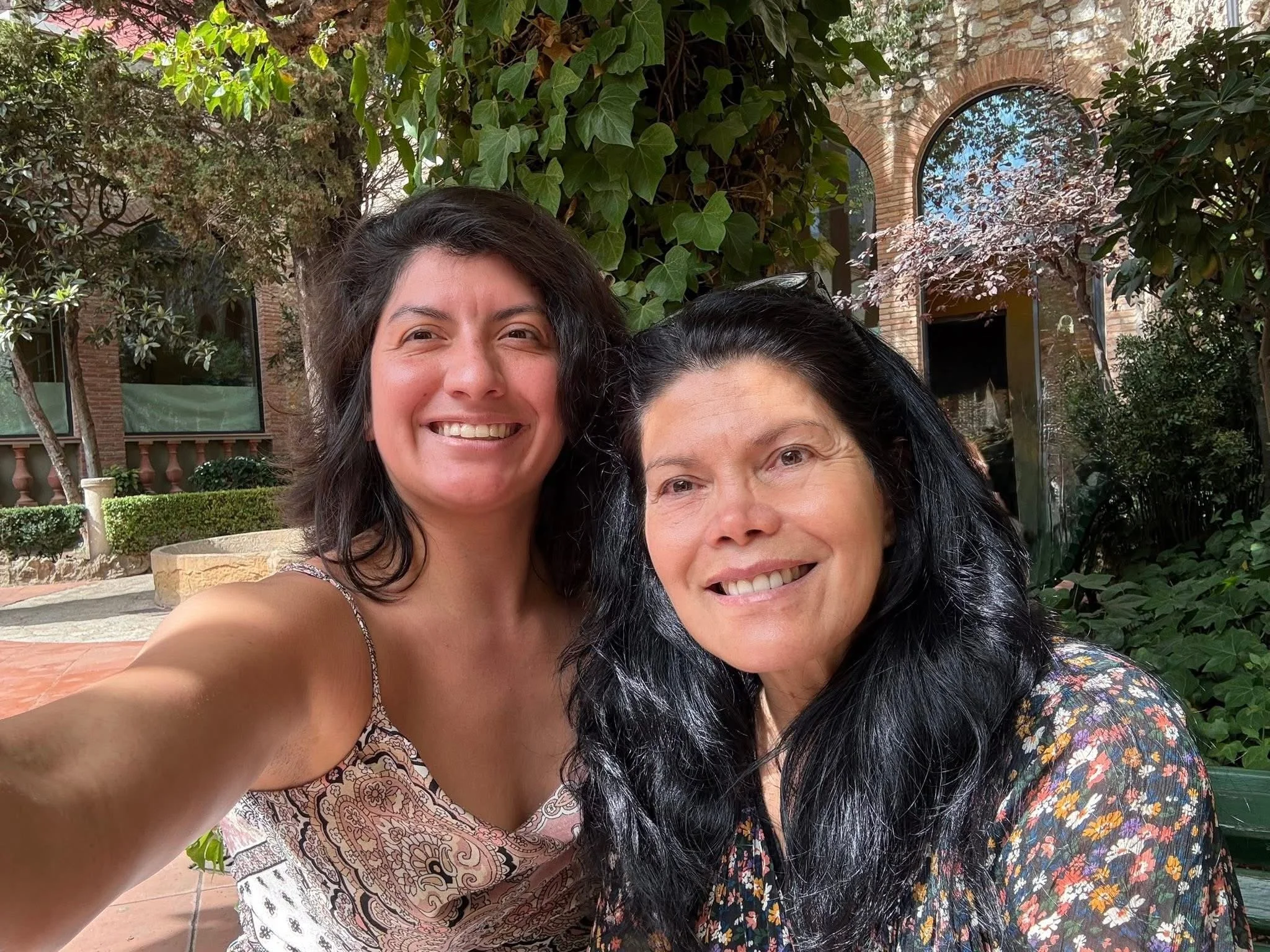 Two women smiling for a selfie outdoors in front of a brick building with arched windows and lush greenery.