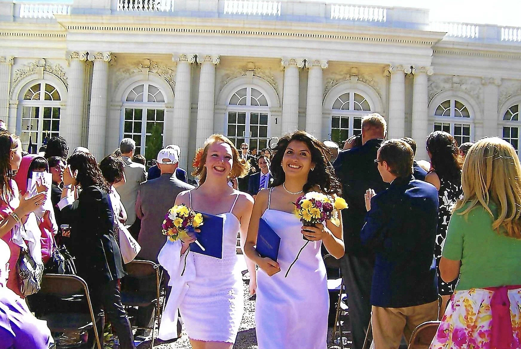 Two women in white dresses holding bouquets walk through a crowd outside a large, ornate white building with tall arched windows and columns, likely during a wedding ceremony.