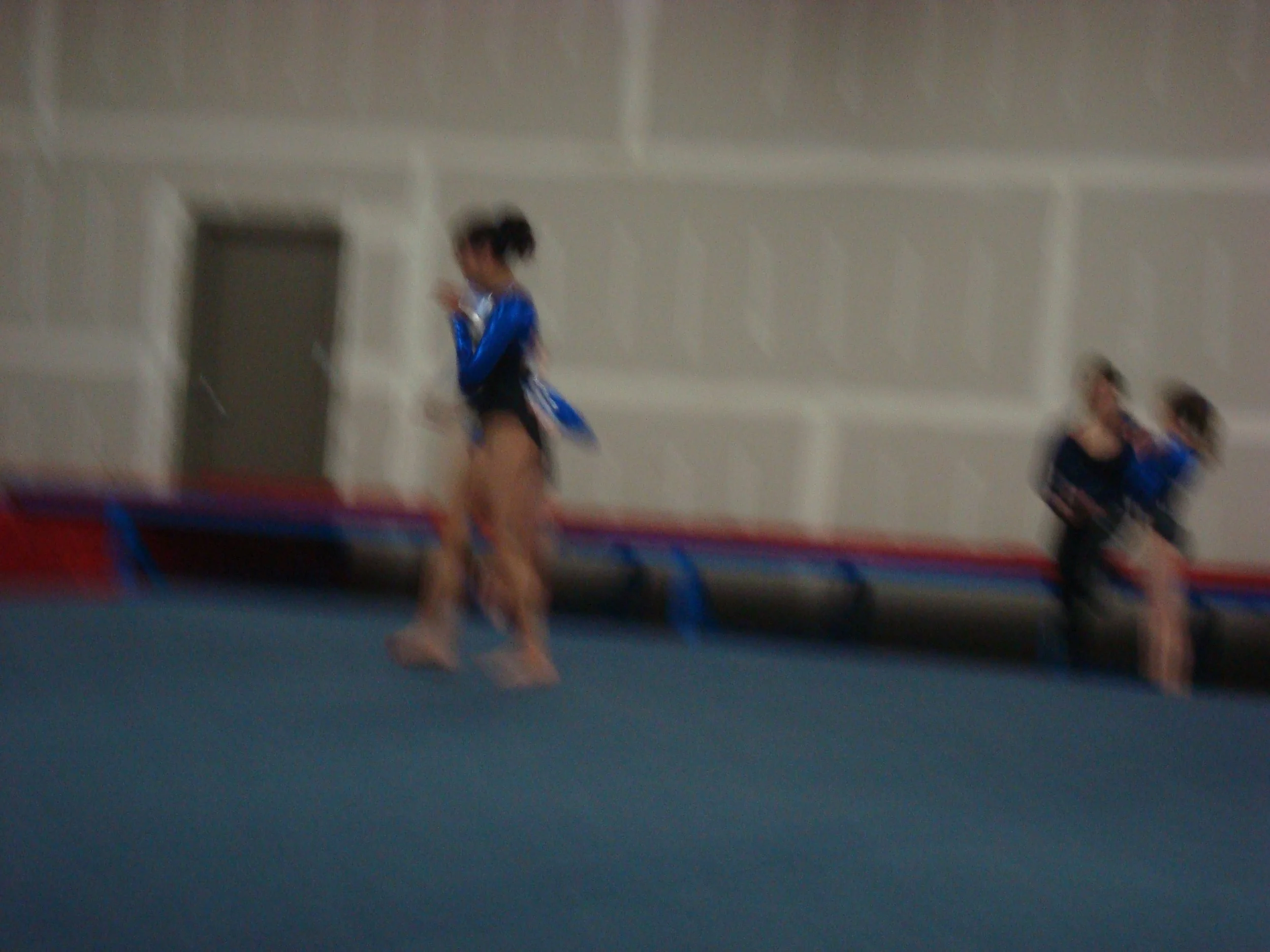 Blurry photo of young girls in bluish athletic outfits practicing gymnastics on mats in an indoor gymnasium.
