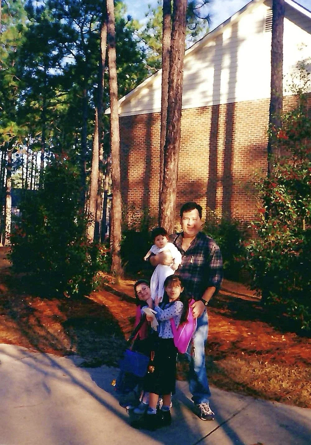 A man with two young girls and a baby standing outdoors in front of a brick building and tall trees, during sunset.