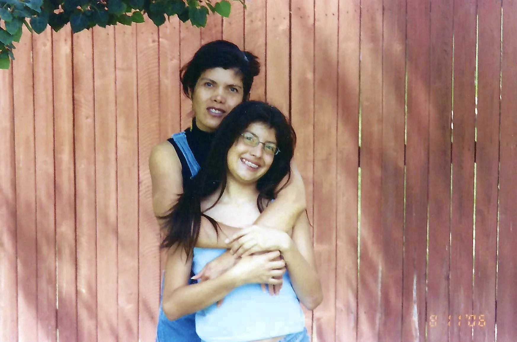 A young woman with long dark hair and glasses stands in front of a wooden fence, smiling, while a person with short dark hair wraps their arms around her from behind, appearing to hug her, in a backyard setting.