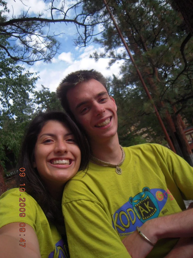 Two smiling young people, a woman and a man, taking a selfie outdoors with trees and a cloudy sky in the background.