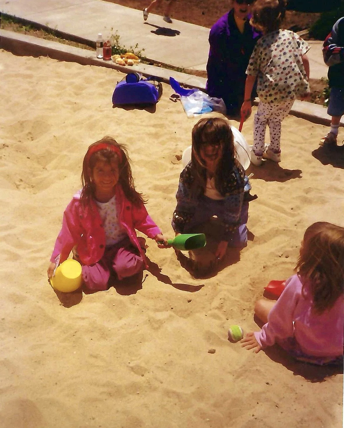 Children playing in the sand, some holding toy shovels and buckets, at a playground or beach area on a sunny day.