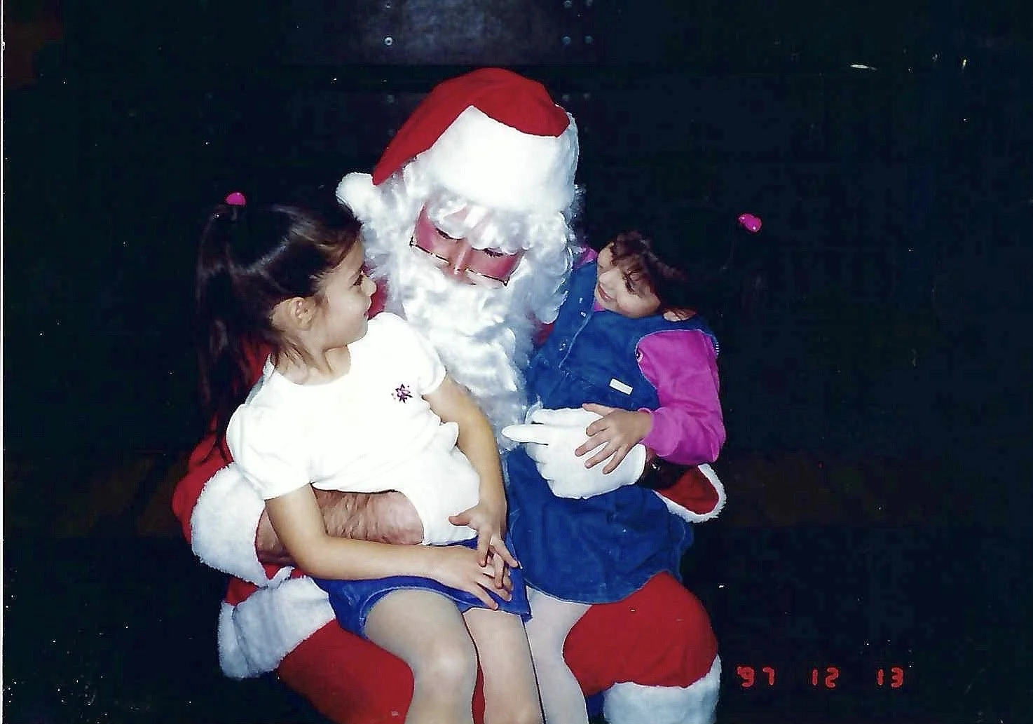 Santa Claus sitting with two young girls, one on each knee, smiling and hugging. Santa is wearing a traditional red suit with white trim, glasses, and a red hat. The girl on the left has dark hair tied back, wearing a white shirt and blue shorts. The