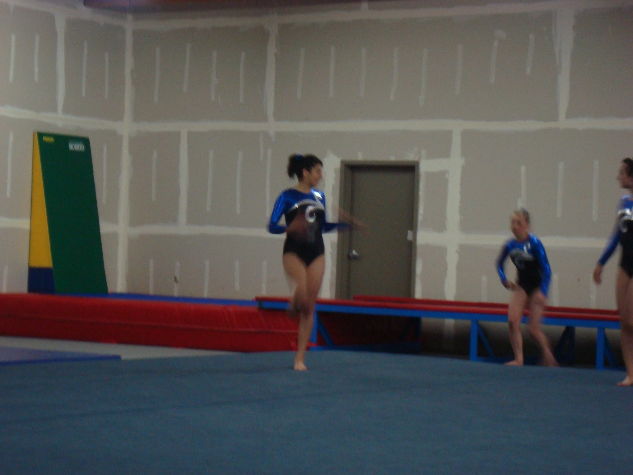 Indoor gym with a trampoline, two female gymnasts in blue uniforms practicing, one standing on the trampoline and the other sitting on a blue mat.