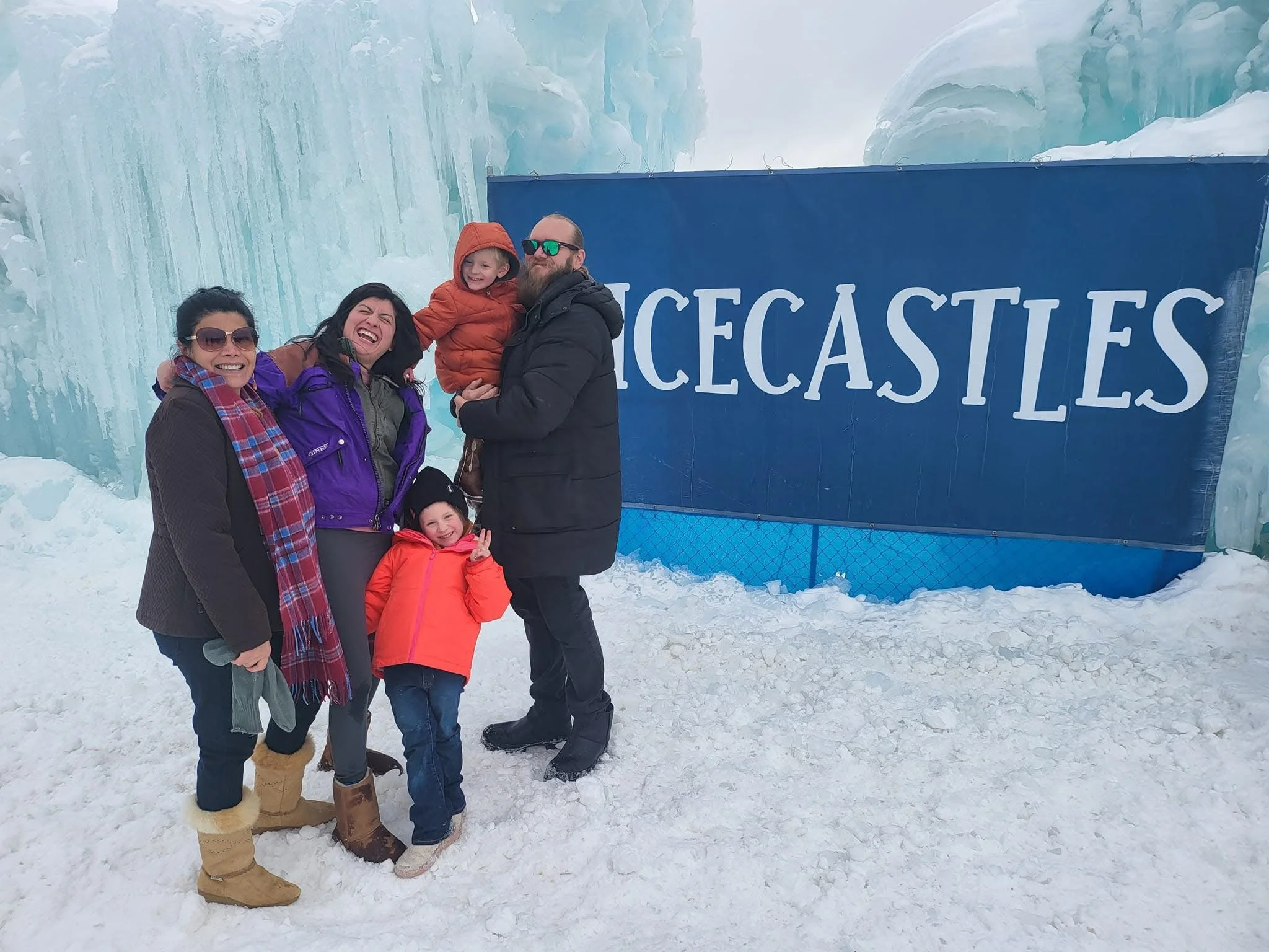 A group of five people, including three children and two adults, standing on snow in front of a large ice formation and a blue sign that reads 'GLACIALS'. They are dressed warmly with jackets, boots, and sunglasses.