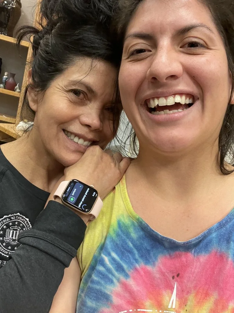 Two women smiling and posing for a selfie. One woman is wearing a tie-dye shirt, and the other has a smartwatch on her wrist.