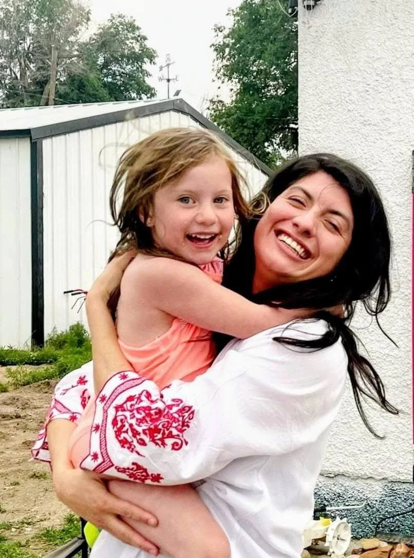 A woman with black hair holding a young girl with light brown hair in her arms, both smiling and hugging outdoors.