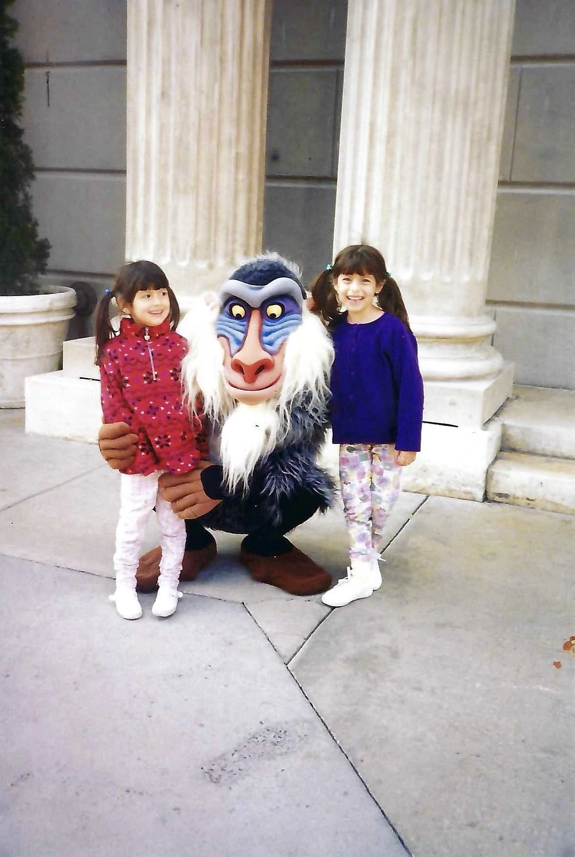 Two young girls posing with a character dressed as Rafiki from The Lion King in front of a classical column structure.