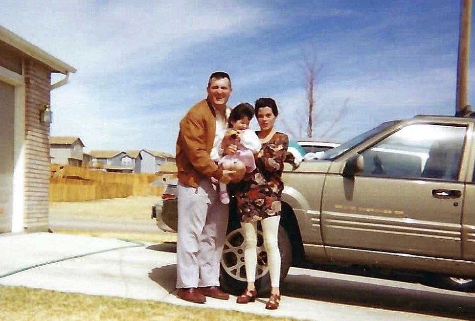 A man, woman, and two children outside in a residential neighborhood, standing next to a silver truck. The man is holding a little girl, all people are smiling.