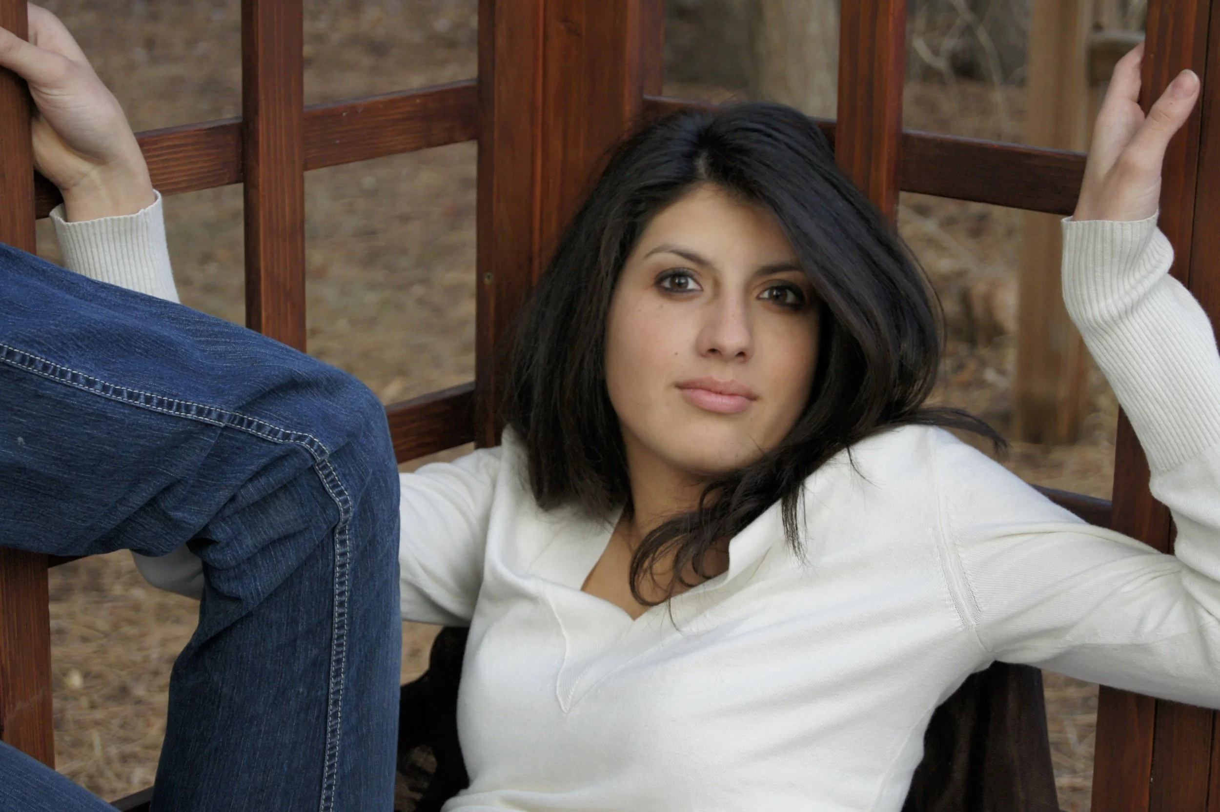 A young woman with dark hair and a white sweater lies on the ground beside a wooden bench, looking at the camera with a neutral expression. Part of a person in jeans is visible, sitting on the bench.