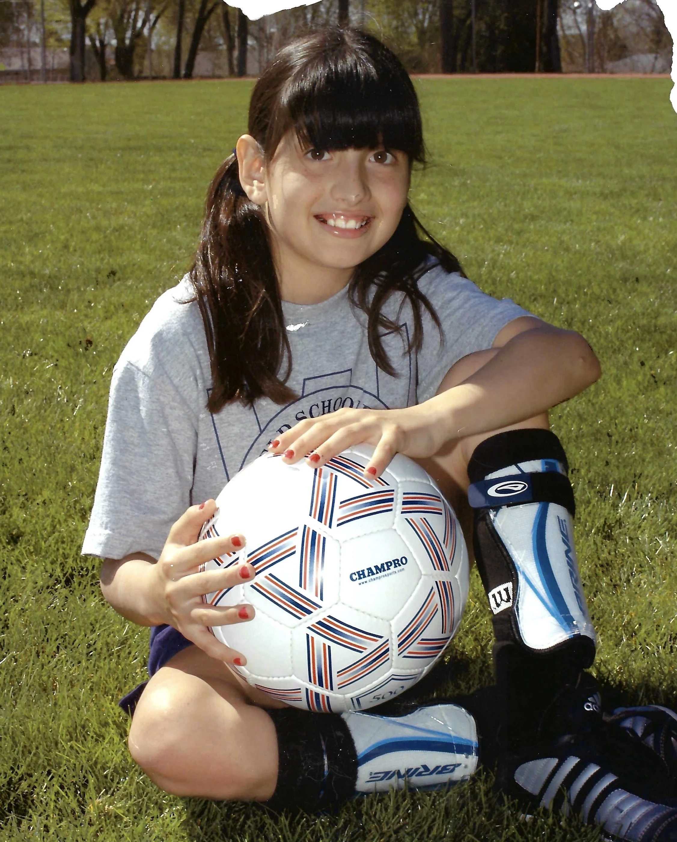 A young girl sitting on grass outdoors, holding a white soccer ball with blue and orange accents, with a water bottle and sports gear beside her.