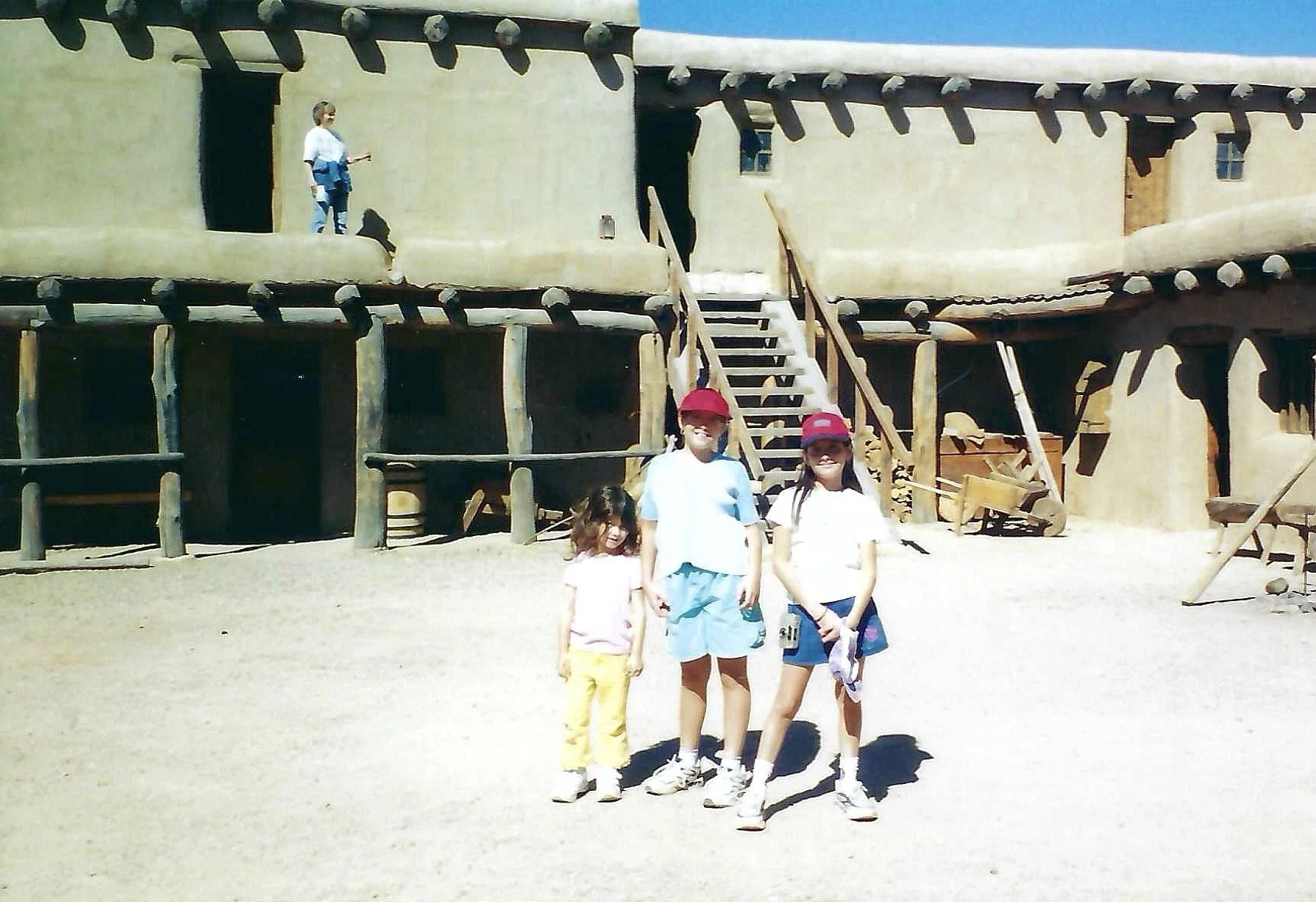 A group of four children standing on a dirt ground in front of a wooden, reproduction adobe-style house with ladders and tools nearby, at a historical site or outdoor museum.