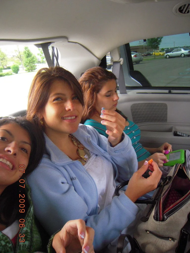 Three young women sitting in the back of a vehicle, with the interior visible and a parking lot seen through the window. The woman in the middle is smiling and holding a small item, while the woman on the right is looking down at a green device, and 