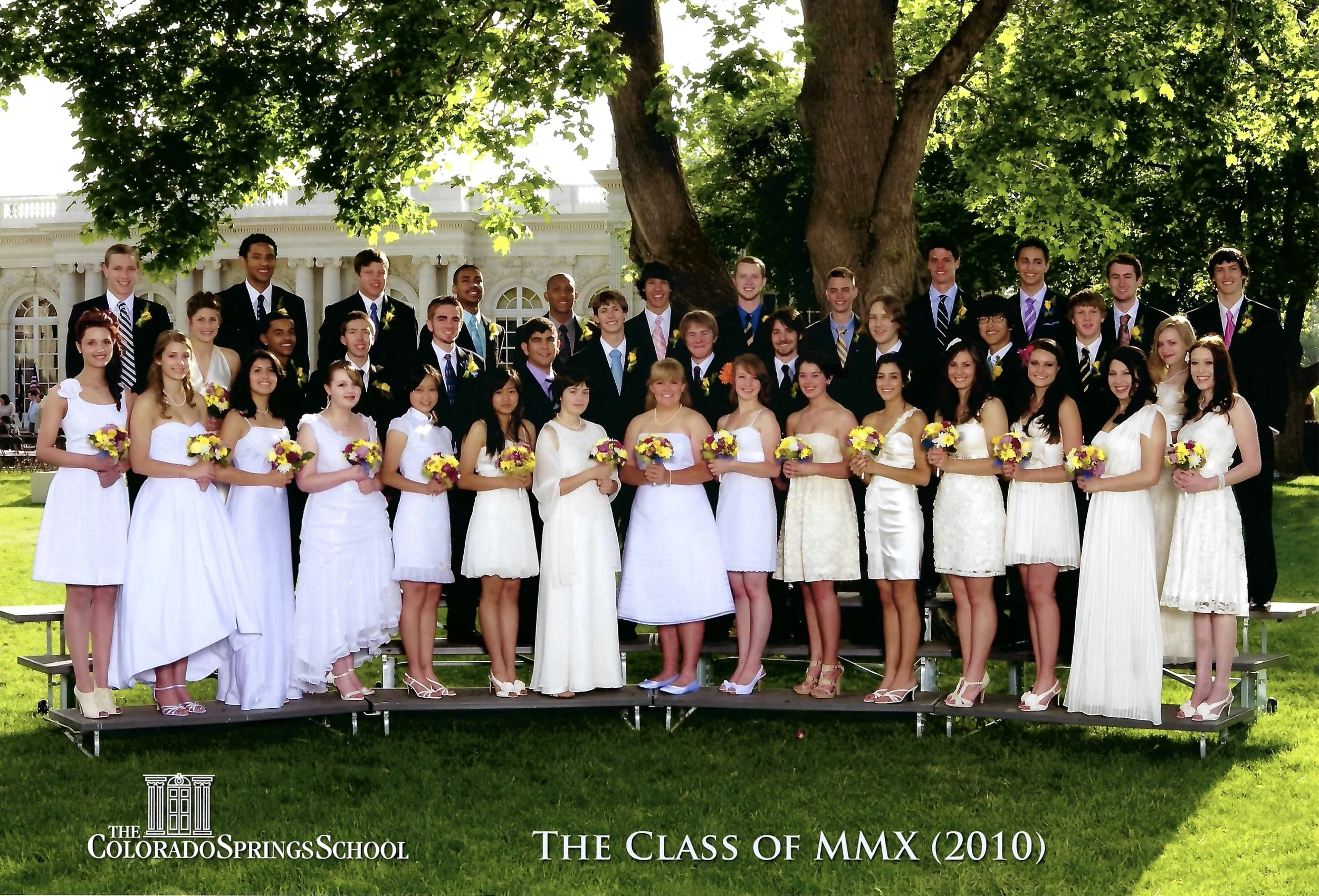 Group of graduates in formal attire, posing outdoors under a large tree, holding yellow and purple flowers, with a building in the background. The photo is from the Class of MMX (2010) at The Colorado Springs School.