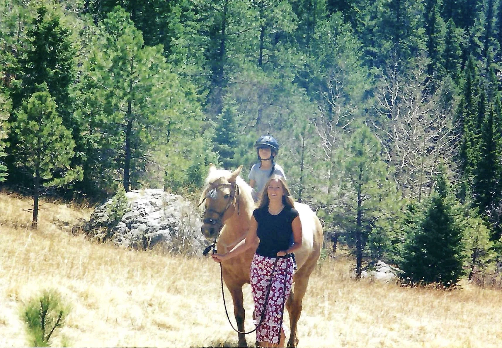A woman standing next to a brown horse with a rider on the horse, in a forested area with tall pine trees and rocky terrain, under a sunny sky.