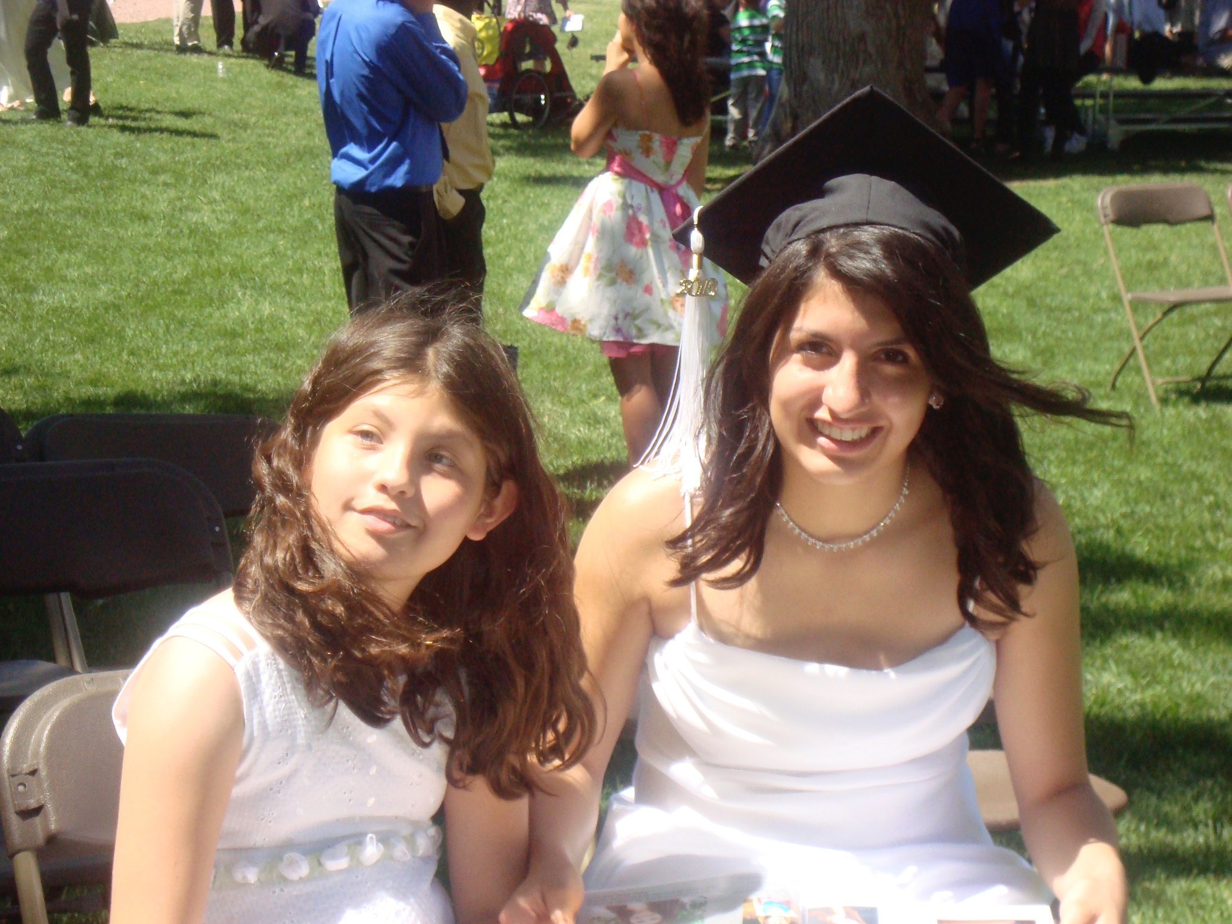 Two young women wearing white dresses sitting outdoors, with one woman wearing a black graduation cap and gown, smiling at the camera. In the background, other people are standing and talking on a grassy area at an outdoor event.