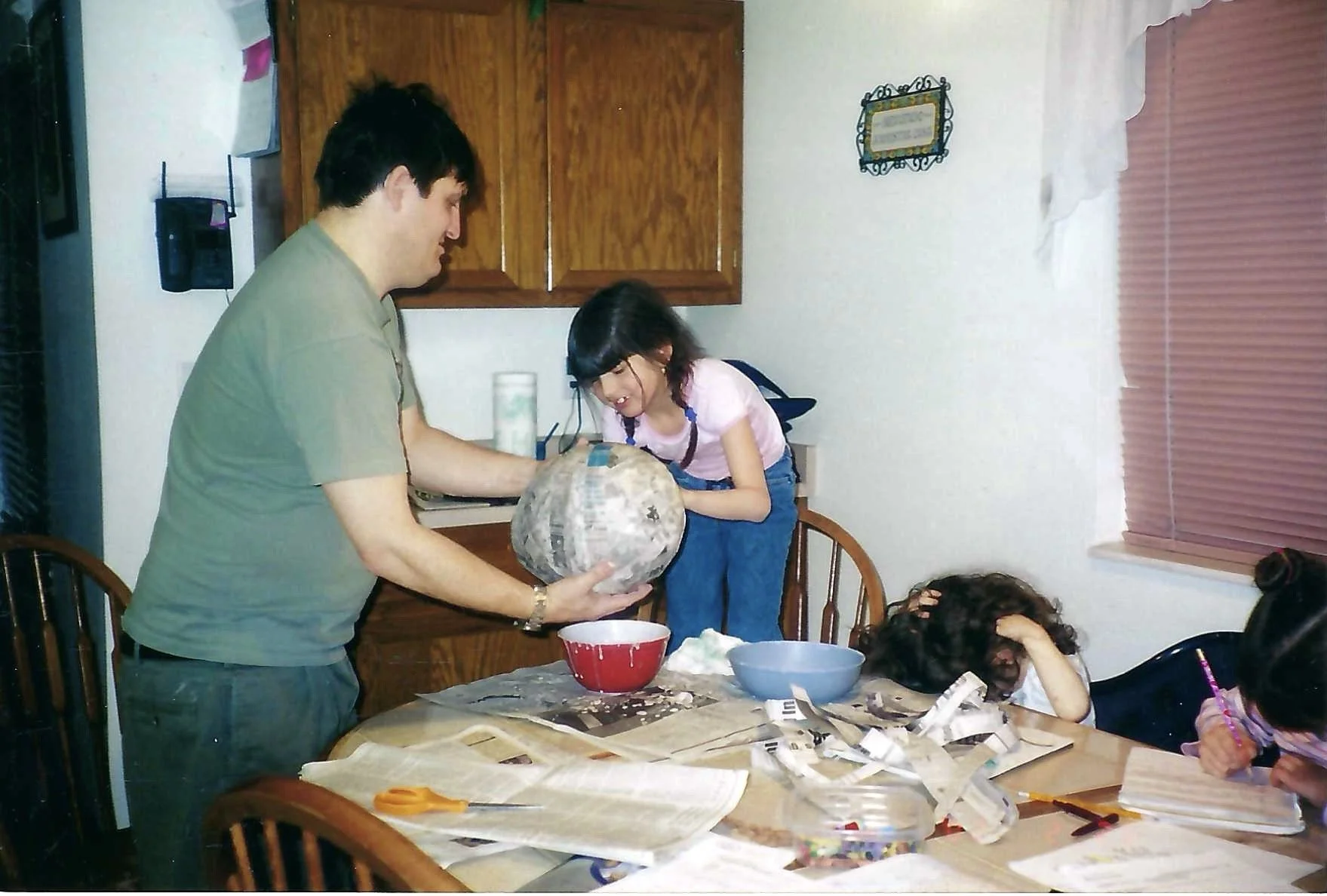 A man is giving a gift to a girl at a family gathering, with another girl sitting at the table looking distressed.