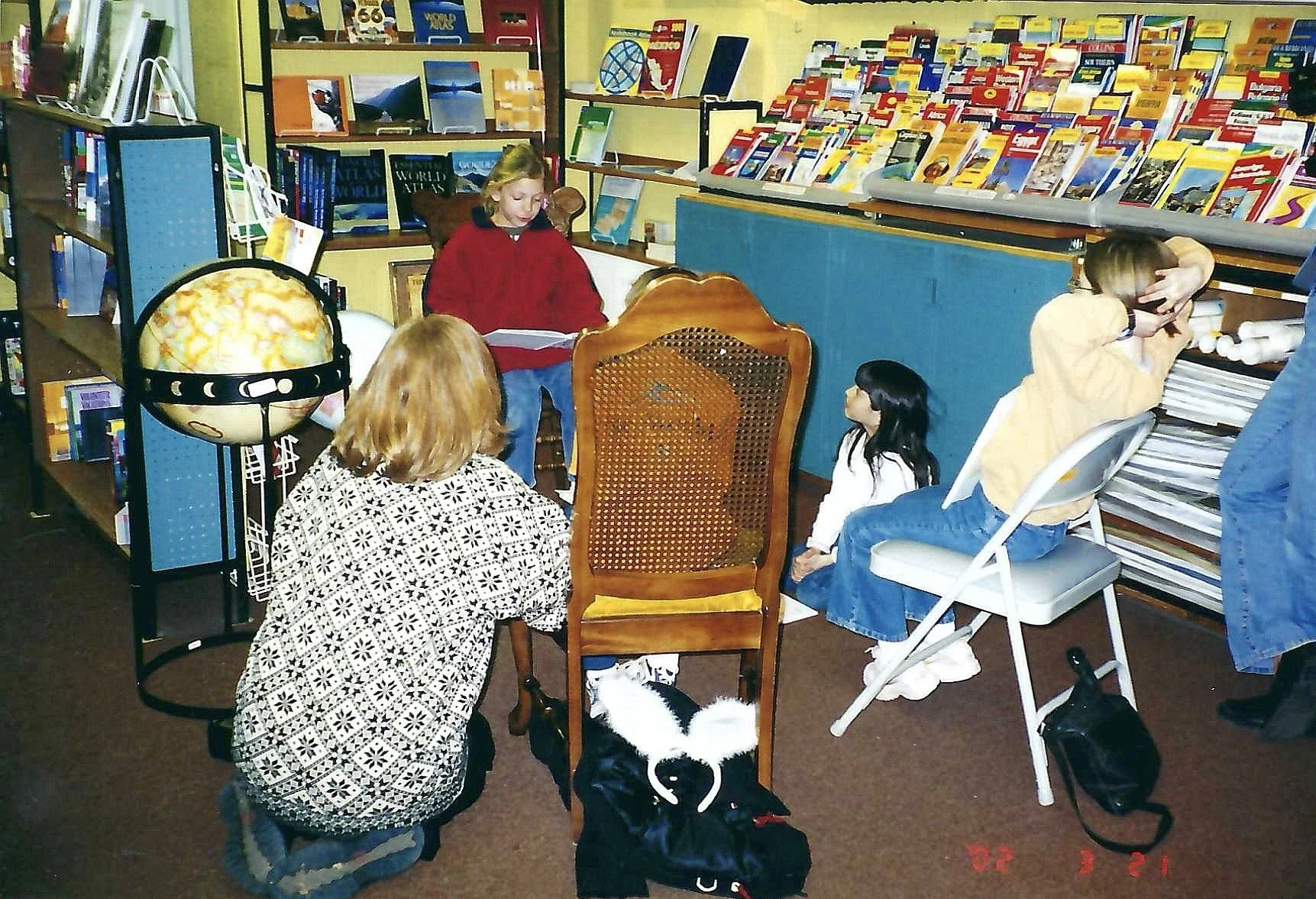 A group of small children and an adult in a bookstore or library, some reading or looking at books, with bookshelves filled with magazines and books in the background. The children are sitting or squatting on the floor, and there is a globe on a stan