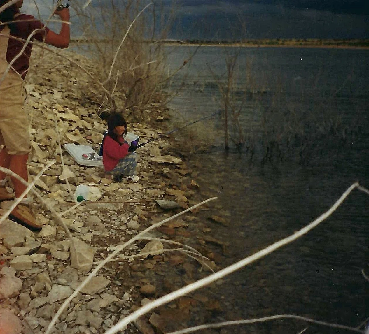 A young girl fishing by a rocky riverside at dusk, with a man standing nearby.