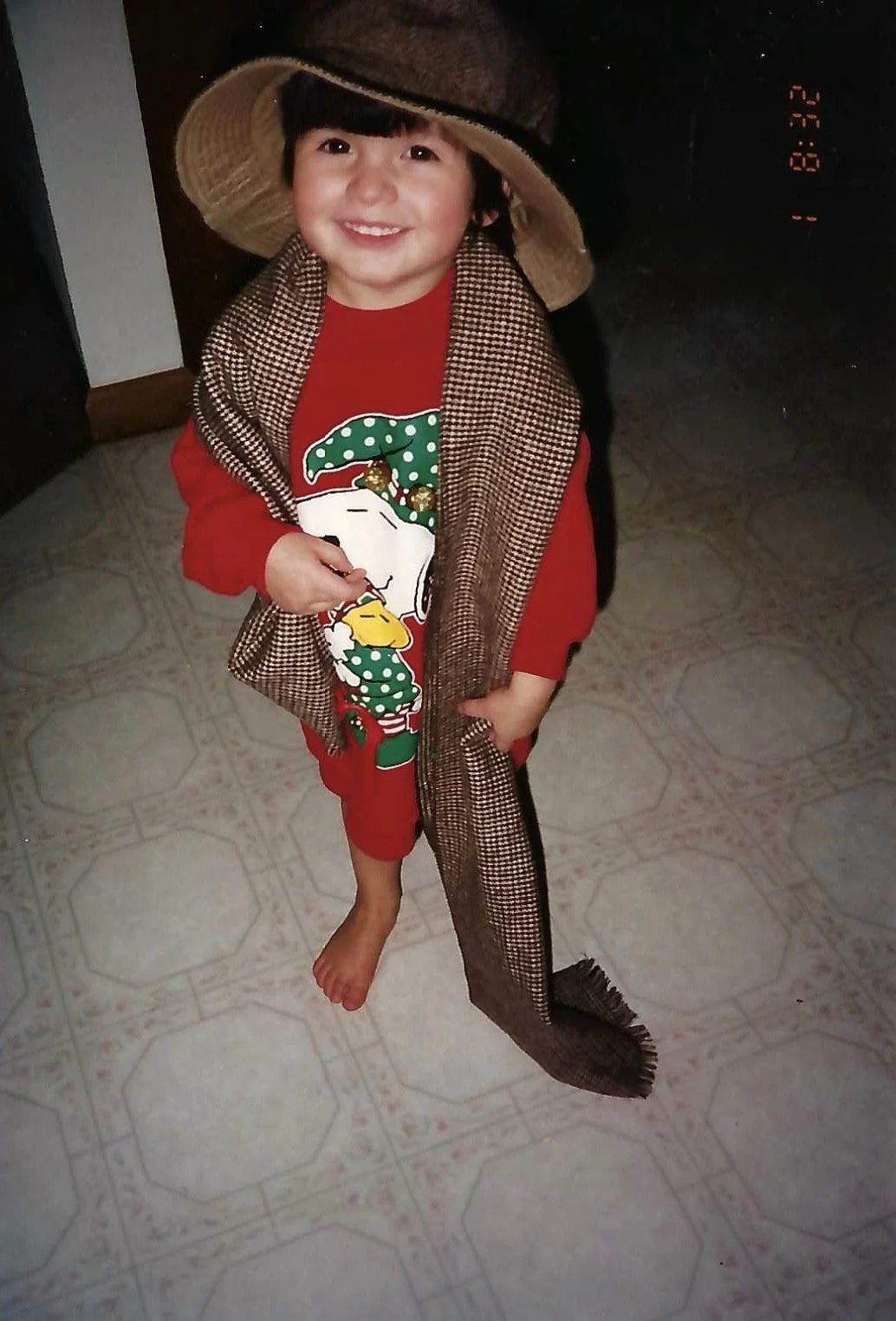 A young boy dressed in holiday themed clothes, wearing a large sun hat, and smiling at the camera. He is standing on a patterned tiled floor, holding a shiny object in one hand, with a long scarf draped around his shoulders.