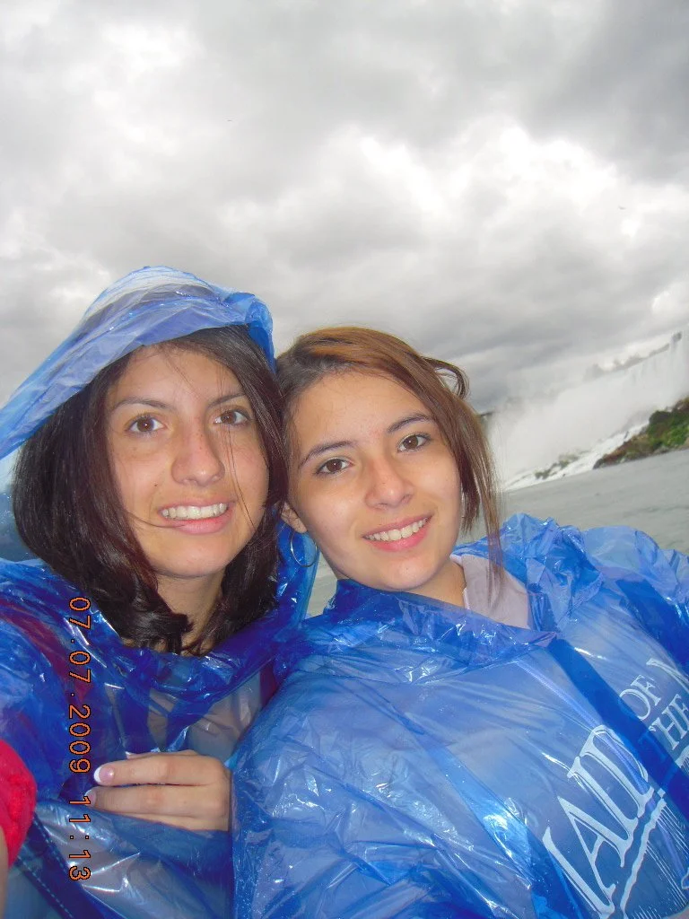 Two young women in blue rain ponchos taking a selfie at Niagara Falls with the waterfall in the background under a cloudy sky.
