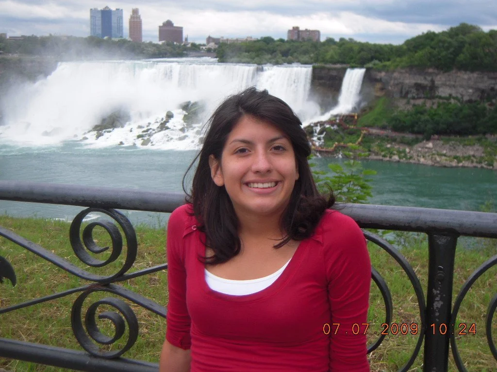 A woman with shoulder-length dark hair smiling in front of Niagara Falls, with a city skyline in the background, wearing a red top and standing by a decorative metal fence on a cloudy day.