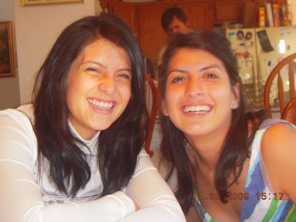 Two women smiling and leaning in together at a table in a kitchen, with a man in the background near a refrigerator decorated with magnets and photos.