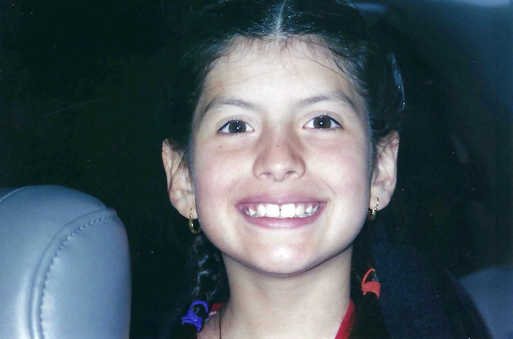 Close-up of a smiling young girl with dark hair, earrings, and a red shirt, lying on her back on a dark surface.