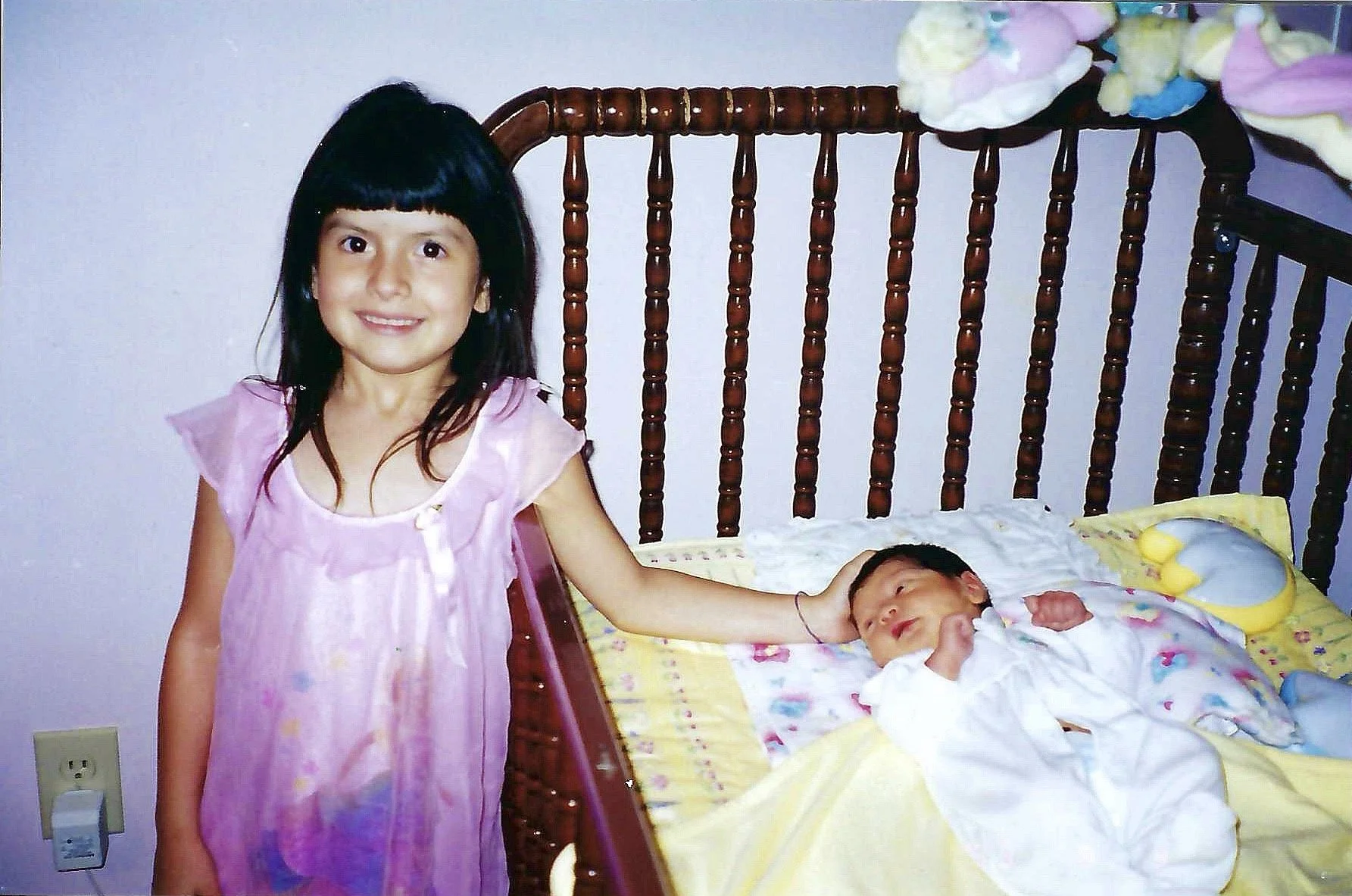 A young girl with black hair and a pink dress standing next to a bed, touching a sleeping baby with her hand.