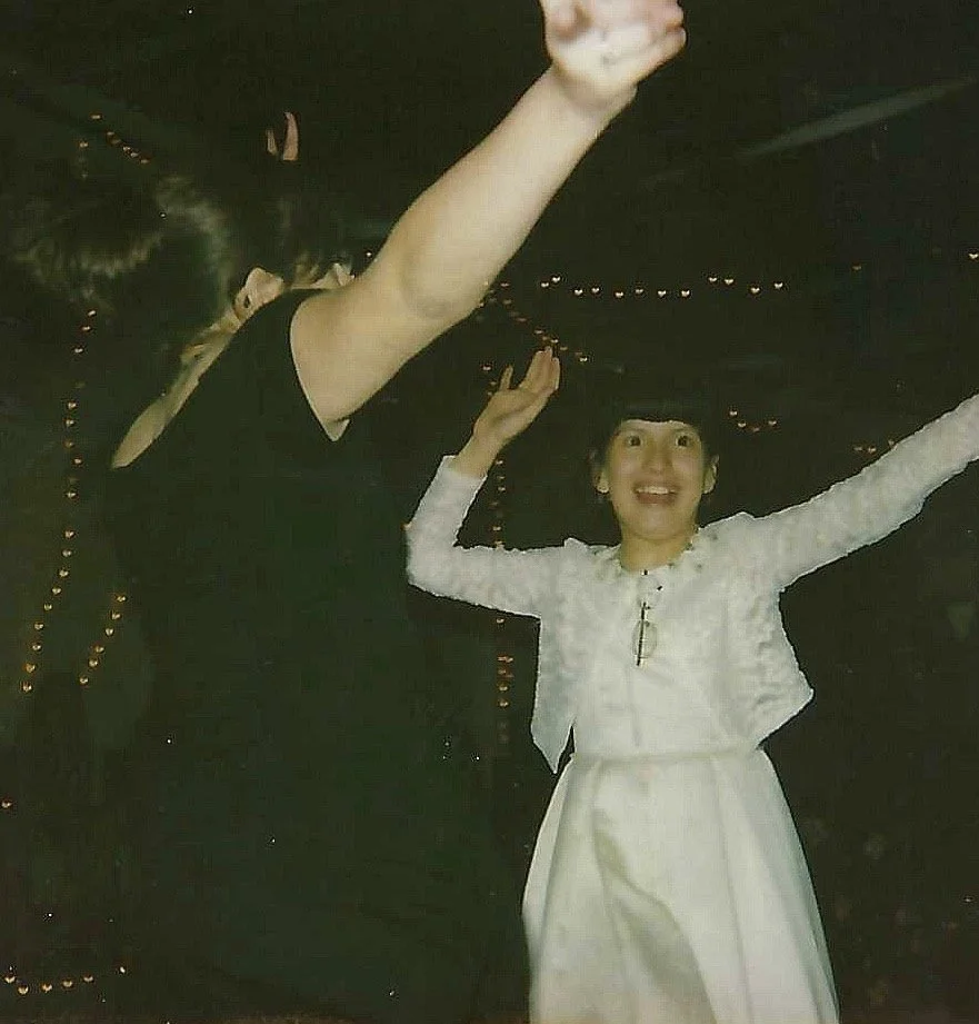 A woman in a white dress with a fitted waist and long sleeves, smiling with her arms raised, appears to be celebrating. The background is dark with small string lights, suggesting an evening or indoor event.
