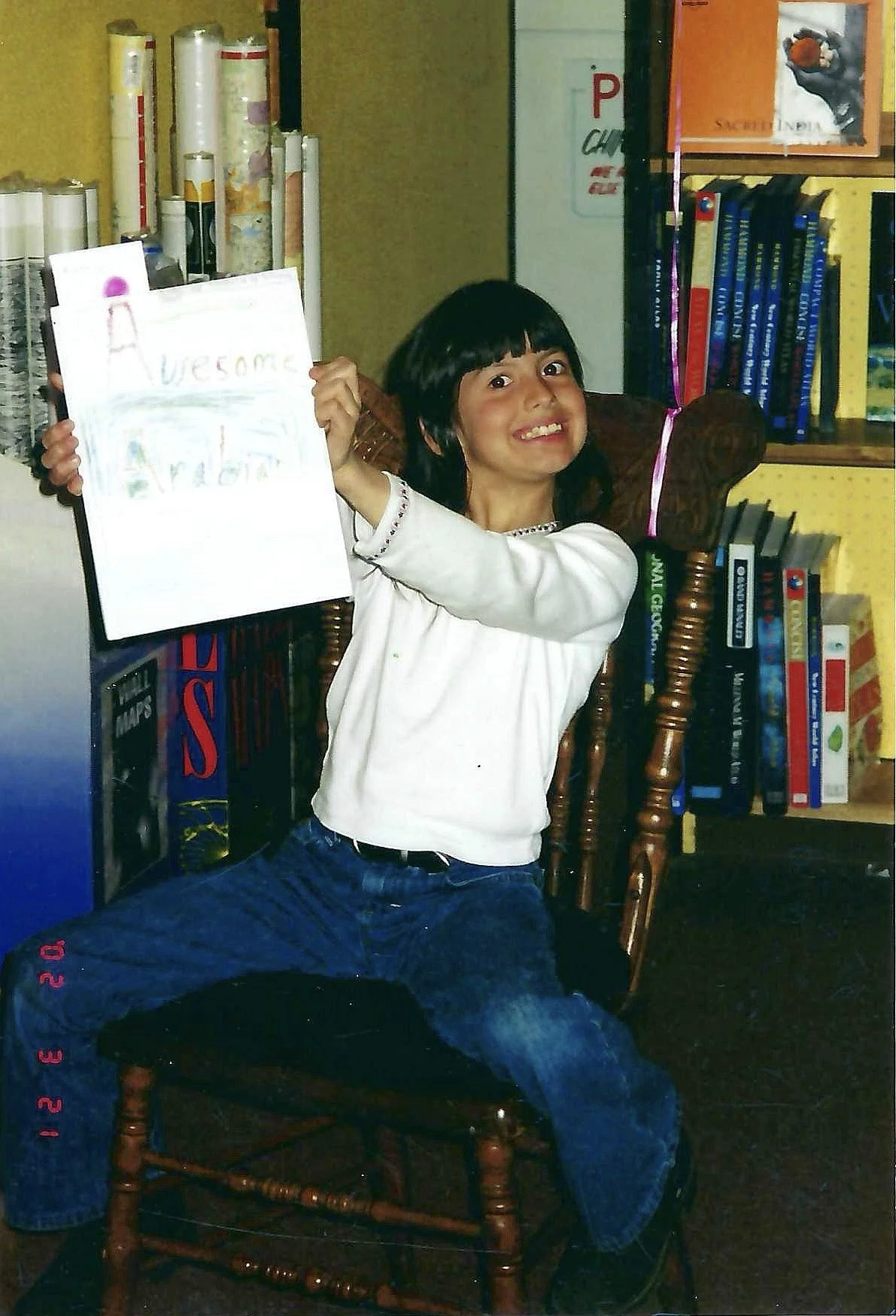 A young girl with black hair and a white long-sleeve shirt is sitting on a wooden chair, holding a colorful drawing that says 'Welcome' and has drawings on it. She is smiling and posing indoors in front of a yellow bookshelf filled with books and dec