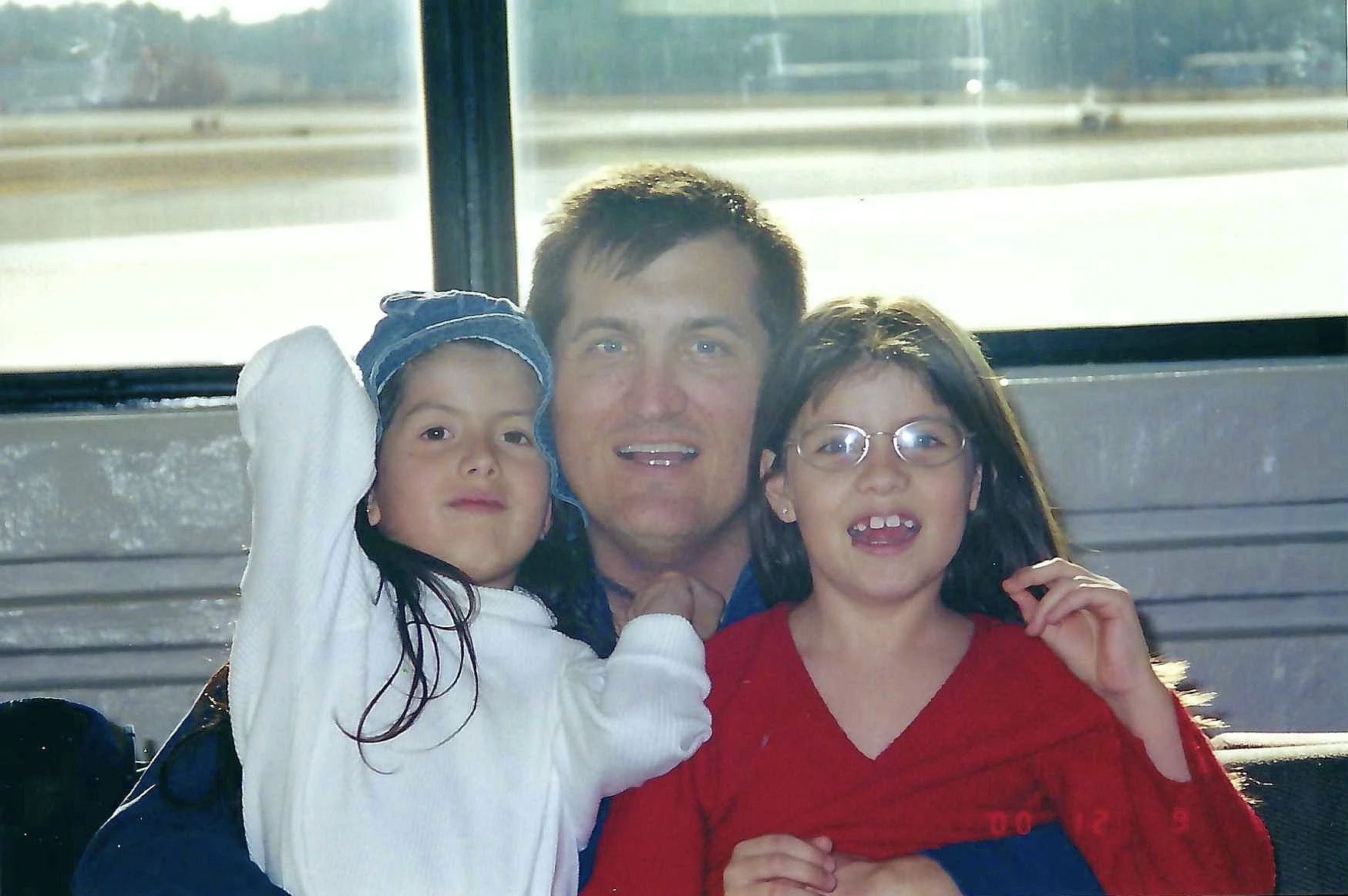 A man sits with two young girls, all smiling, on a bus with a window showing a highway outside.